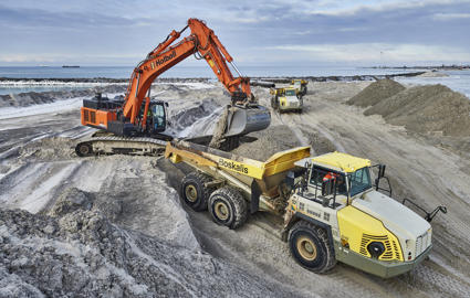 English: An excavator lifts sand into a dumper truck.// Dansk: En gravemaskine løfter sand op i en dumper.//Deutsch: Ein Bagger schüttet Sand in einen Muldenkipper.