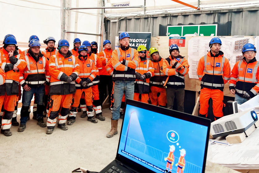 En gruppe arbejdere i orange arbejdstøj og blå hjelme får sikkerhedsuddannelse. I forgrunden ses en skærm med en forklarende video.