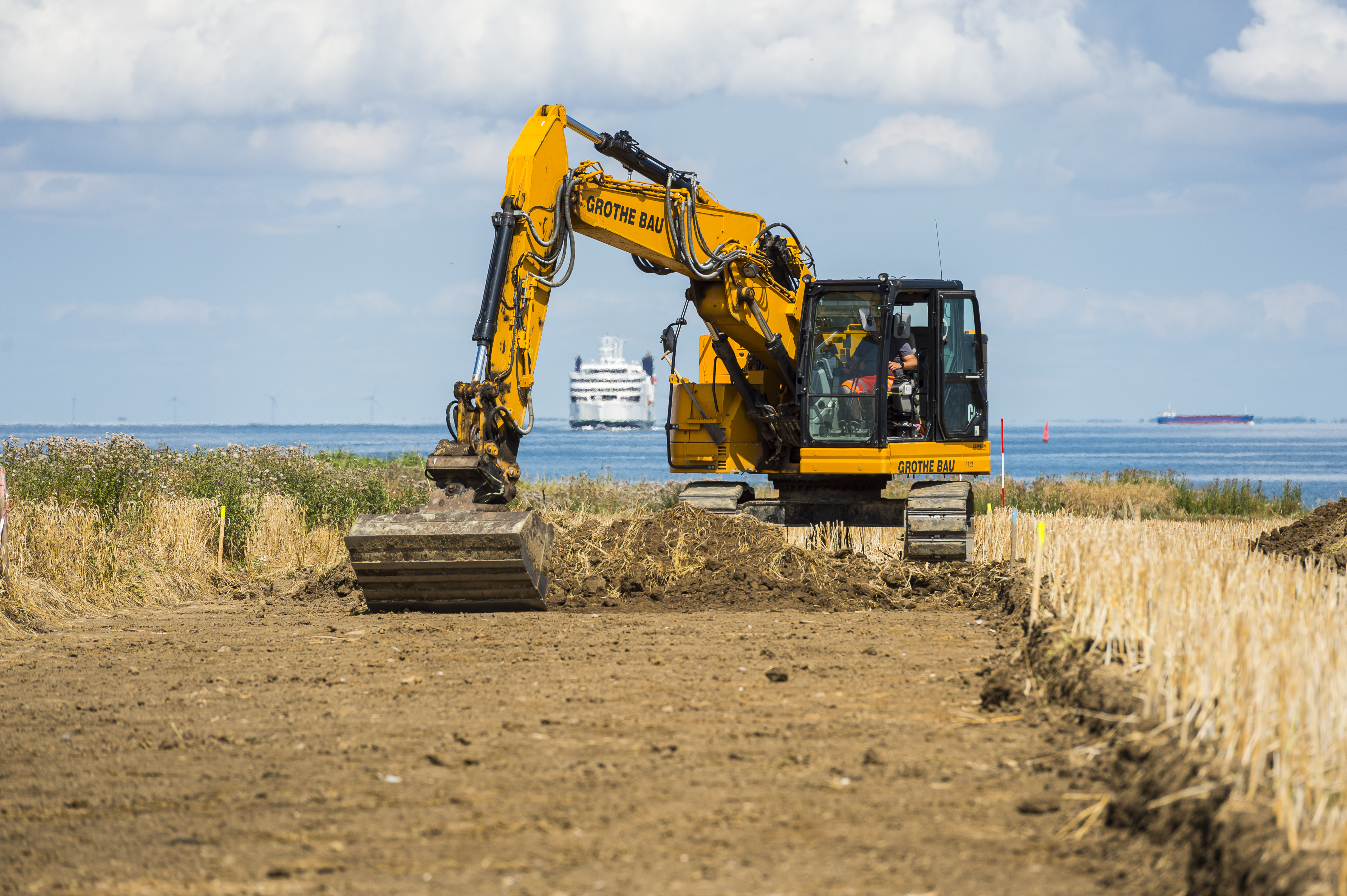 Ein gelber Bagger arbeitet auf der Baustelle. Im Hintergrund die Ostsee.