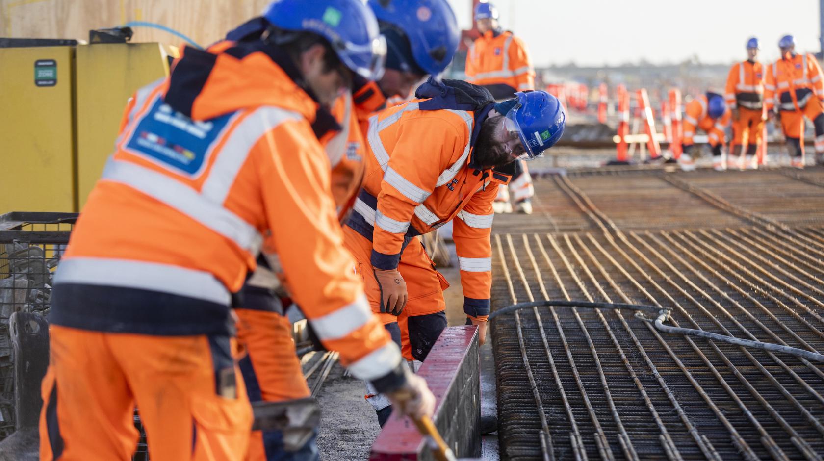 Arbeiter in orangefarbener Arbeitskleidung arbeiten mit Bewehrungsstahl.