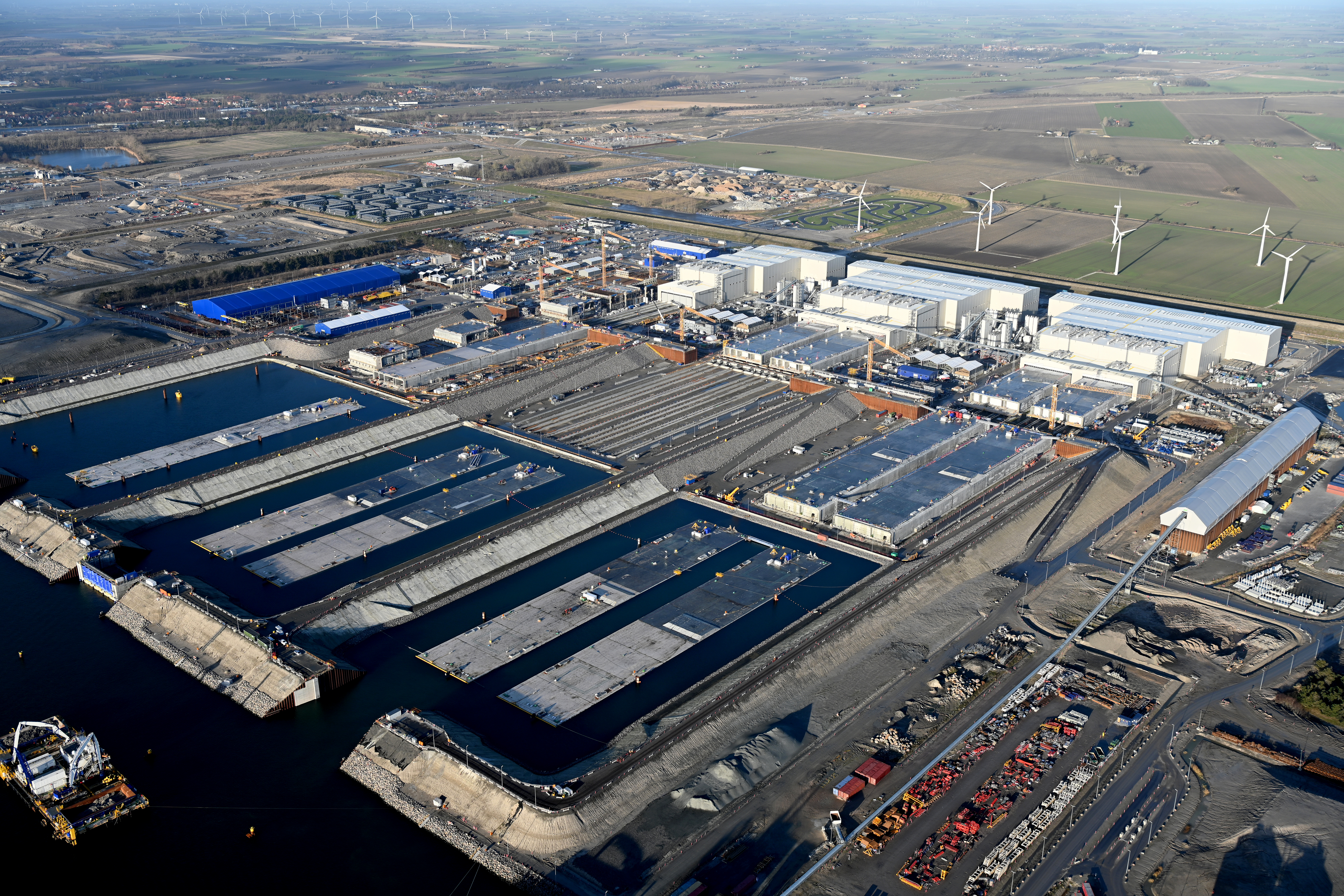 The tunnel element factory near Rødbyhavn. You can see the three large white factory buildings where the tunnel elements are manufactured. Finished cast elements lie in front of the factory buildings. Some are already floating in the water.