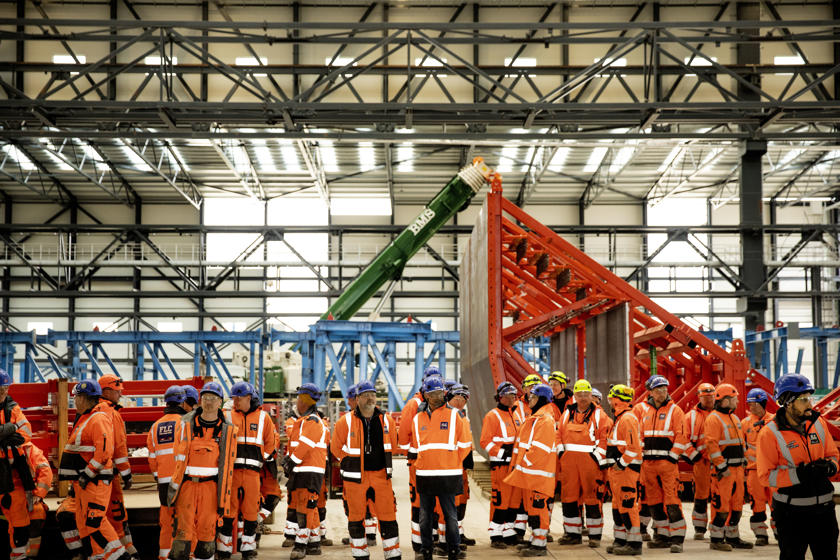Mange personer i orange arbejdstøj og med blå og gule hjelme står i en fabrikshal.