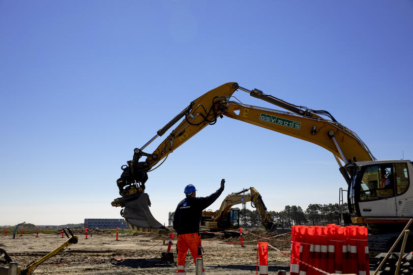 Ein gelber Bagger bei der Arbeit auf der Baustelle. Im Vordergrund steht ein Arbeiter mit Helm, der dem Baggerfahrer Zeichen gibt. Im Hintergrund ist ein weiterer gelber Bagger zu sehen.