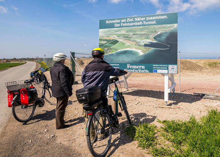 EN: Two cyclists standing in front of an information sign next to the construction site. // DE: Zwei Fahrradfahrer stehen vor einem Informationsschild neben der Baustelle. // DA: To cyklister står foran et informationsskilt ved siden af byggepladsen.
