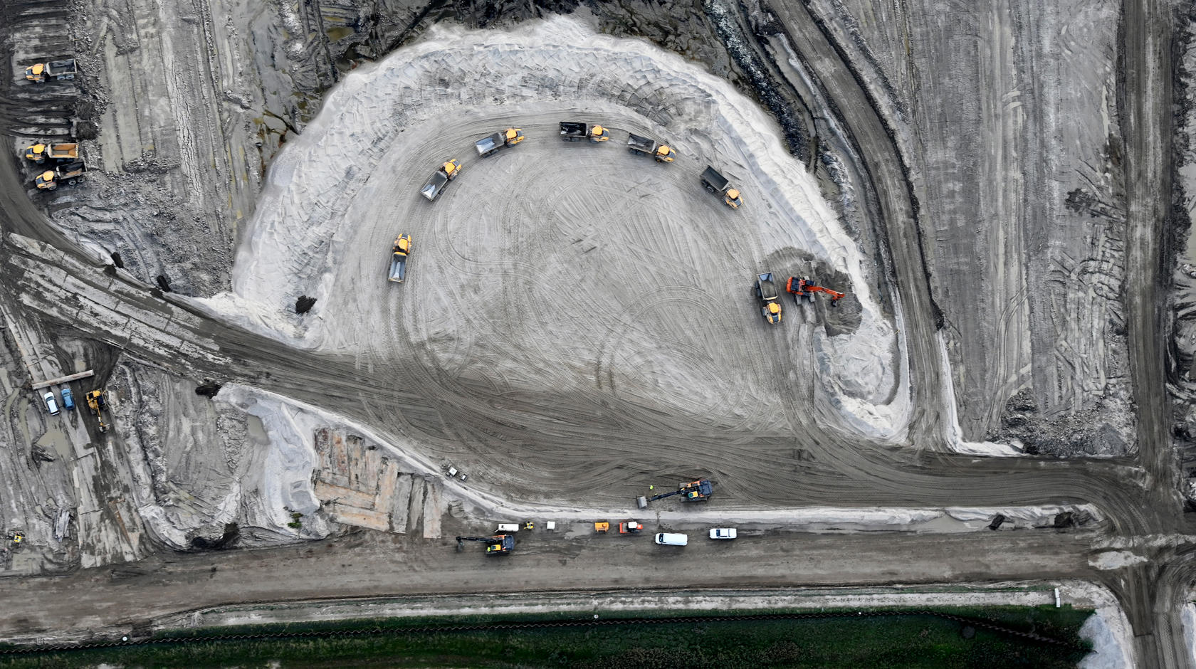 Blick auf eine Sandfläche auf der Baustelle von oben. Im Halbkreis stehen Dumper an, um von einem Bagger mit Material beladen zu werden.