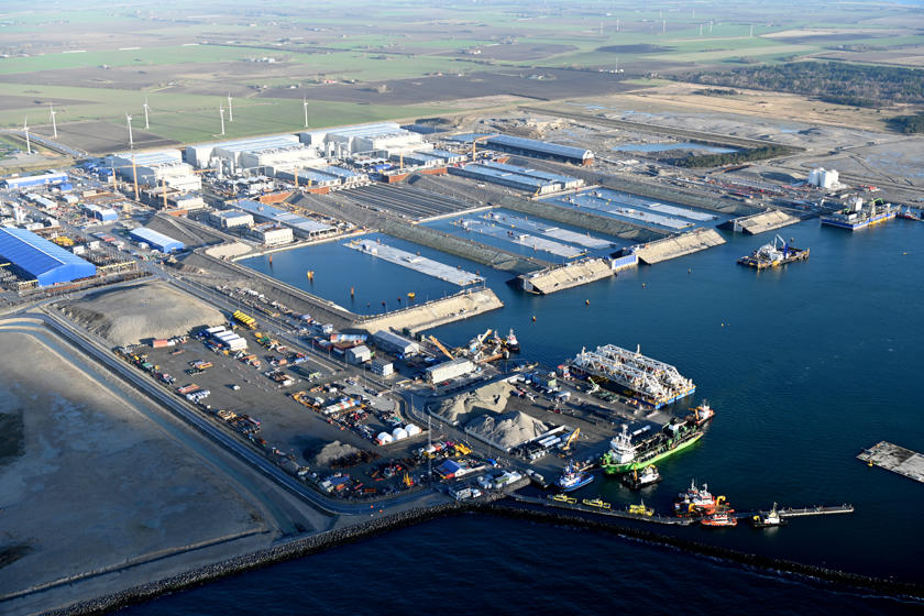 Die Tunnelfabrik auf Lolland. Große Tunnelelemente aus Beton werden in drei großen weißen Hallen produziert. Vor den Hallen sind Wasserbecken, über die die Elemente ausgeschwommen werden. Einige Elemente schwimmen im Wasser.