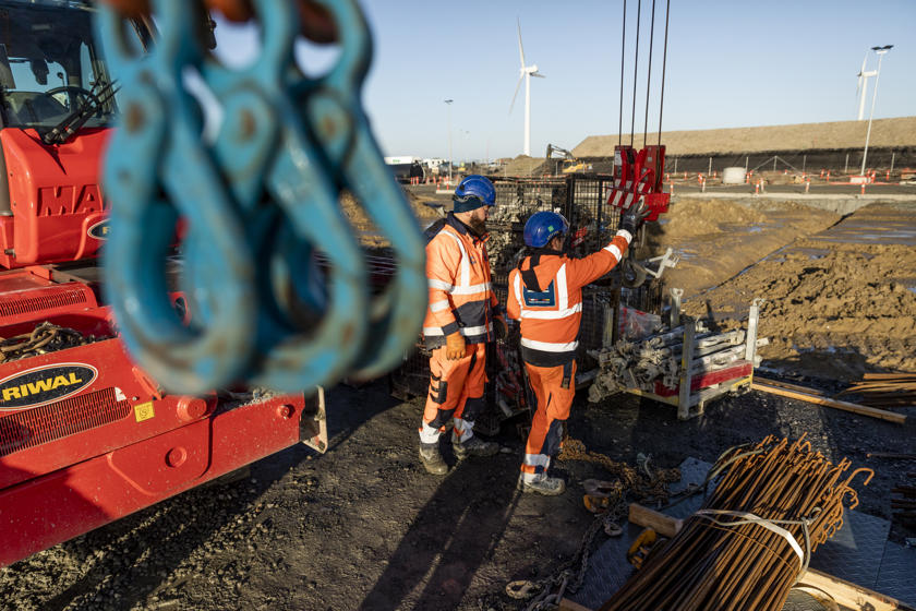 To arbejdere i orange arbejdstøj og blå hjelme arbejder på byggepladsen med en kran.