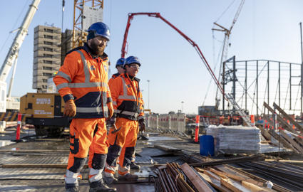 Tre mænd i orange arbejdstøj står på byggepladsen. I baggrunden står en kran. Foran dem ligger armeringsjern.