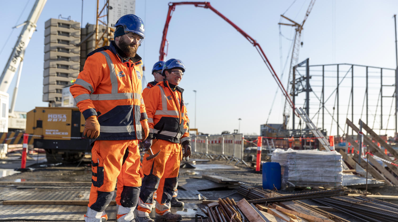 Tre mænd i orange arbejdstøj står på byggepladsen. I baggrunden står en kran. Foran dem ligger armeringsjern.