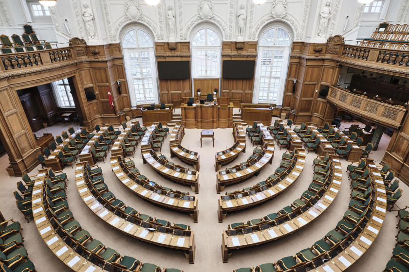 The Folketing's chamber. The walls are panelled with wood. The tables and chairs are arranged in a semicircle. A lectern stands in the centre.