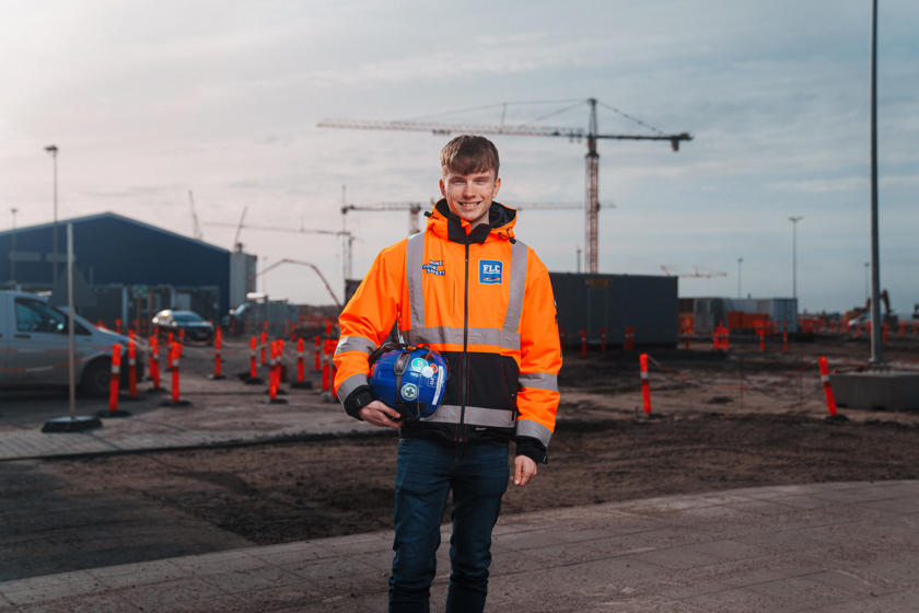 En ung mand i orange arbejdsjakke står foran byggepladsen med en blå hjelm i hånden. I baggrunden ses arbejdskraner.