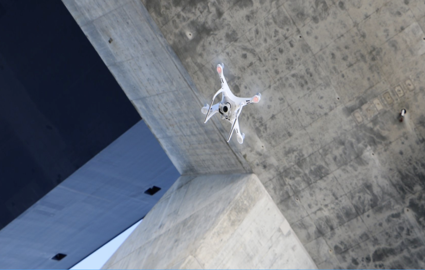 Blick von unten auf eine fliegende Drohne. Im Hintergrund ist der Beton eines Brückenpfeilers zu sehen.