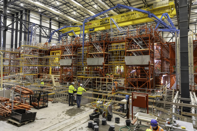 Blick auf die Schalungen für die Betonage eines Tunnelelements in der Tunnelfabrik. Die gelben Schalungen werden von einem roten Gerüst getragen. Blaue Betonpumpen befinden sich über dem Gerüst.